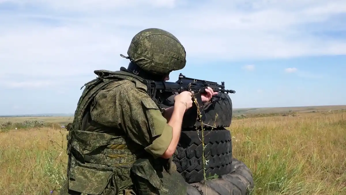 солдат с автоматом в руках в шлеме и бронежилете сидит в поле готовится стрелять учения